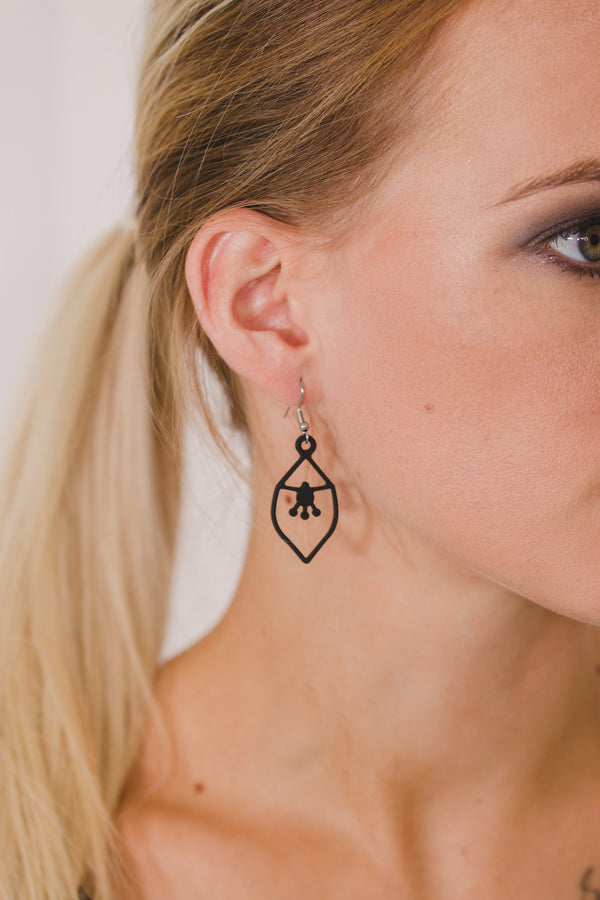 zoom, vue gros plan, boucles d'oreilles Nymphéa, couleur noir, portées sur mannequin Dorine. Attache dormeuse en acier inoxydable. Bijoux fantaisies, marque française, créateurs, designers. Petit modèle, style minimaliste, losange, pampilles, fleurs, géométrique. Silicone, souple, léger, minimisant les allergies, résistant à l'eau/à la chaleur/à la lumière. Idée cadeau original.